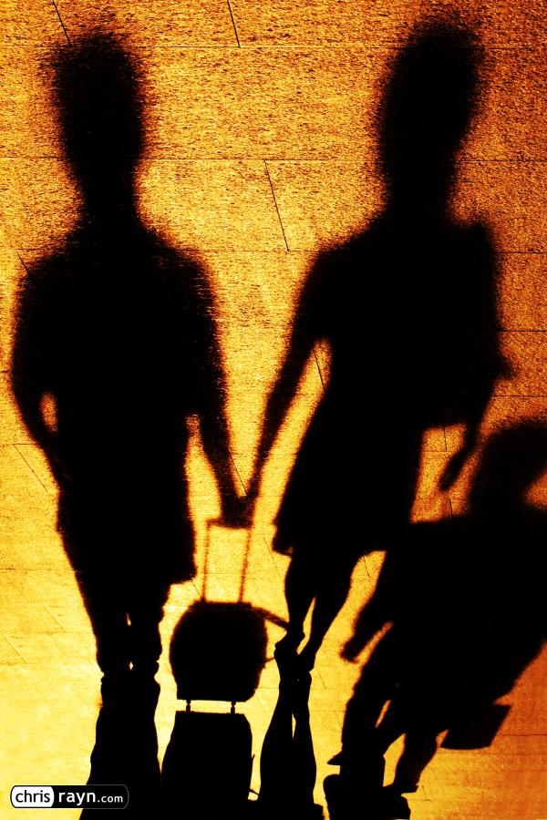 A couple carries luggage together, their shadows in cooperat